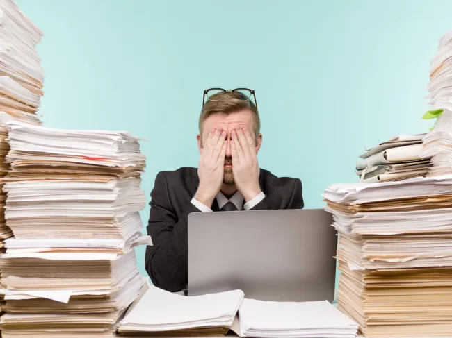 Man covering his eyes, surrounded by stacks of papers